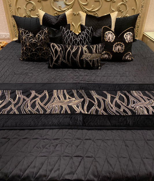 A black bedding set with ornate gold cushions and bed cover, displayed on a bed with a decorative bed frame in the background.